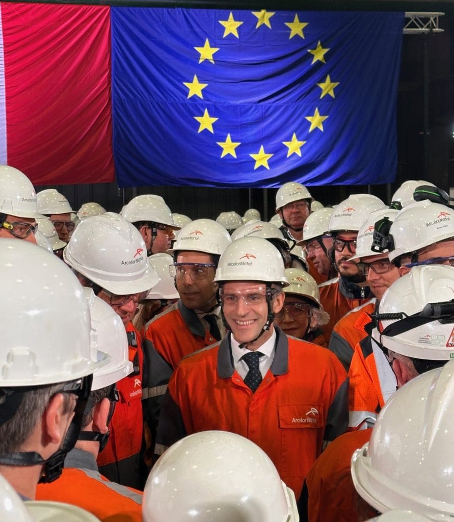 Visite officielle sur un site industriel ArcelorMittal : échange avec les équipes en tenue de sécurité (combinaison orange, casque blanc), devant les drapeaux français et européen